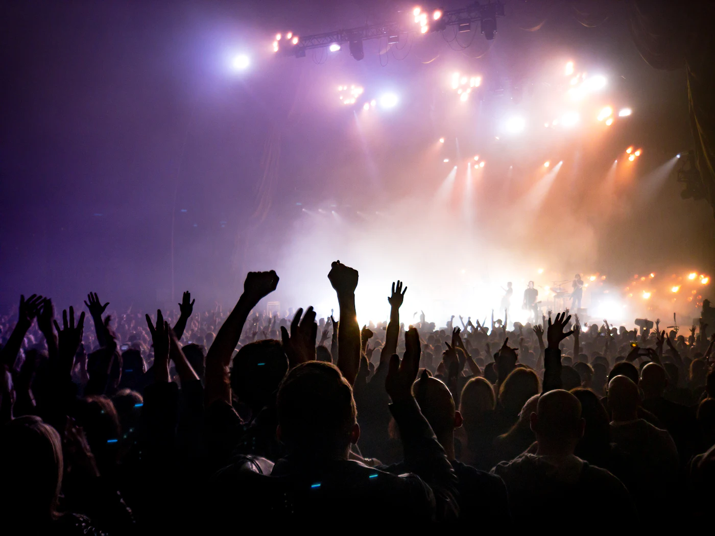 Concertgoers with hands raised as warm lights cut through the stage haze